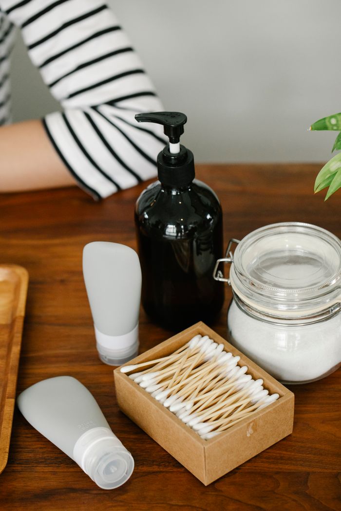 Anonymous person sitting near white plastic tubes of skincare cosmetics lotion dispenser glass jar of white cream and ear sticks in box on table